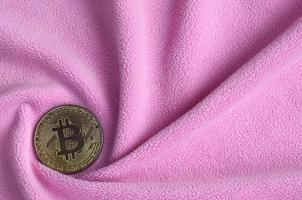 The golden bitcoin lies on a blanket made of soft and fluffy light pink fleece fabric with a large number of relief folds. The shape of the folds resembles a fan from a video card cooler photo