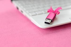 Brilliant pink usb flash memory card with a pink bow lies on a blanket of soft and furry light pink fleece fabric beside to a white laptop. Classic female gift design for a memory card photo