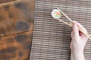 A hand with chopsticks holds a sushi roll on a bamboo straw serwing mat background. Traditional Asian food photo