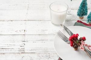 Christmas table setting with white dishware, silverware and red and green decorations on white wooden background. Top view. photo