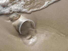 cerrar la vista superior de un vaso de plástico transparente de desecho en la playa de arena, un momento antes de que la ola del mar lo precipitara, con espacio para copiar. foto
