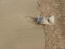 cerrar la vista superior de un vaso de plástico transparente de desecho en la playa de arena, un momento antes de que la ola del mar lo precipitara, con espacio para copiar. foto