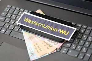 TERNOPIL, UKRAINE - SEPTEMBER 6, 2022 Western Union paper logotype lies on black laptop with ukrainian hryvnias. Western Union Company is American multinational financial services company photo