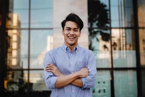 feliz joven empresario de pie con una gran sonrisa en la ciudad. brazos cruzados y mirando a cámara foto