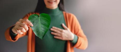 Green Energy, ESG, Renewable and Sustainable Resources. Environmental and Ecology Care Concept. Close up of Smiling Woman Holding a Heart Shape Green Leaf, presenting to Camera photo