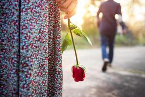 tristeza amor al final del concepto de relación, mujer con el corazón roto de pie con una rosa roja en la mano, hombre borroso en la parte de atrás alejándose como fondo foto