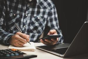 Man holding a phone with a pen and paper to write a note in an office diary using a laptop at office. photo