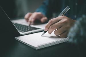 Man hand with pen writing on notebook with computer laptop at desk. photo