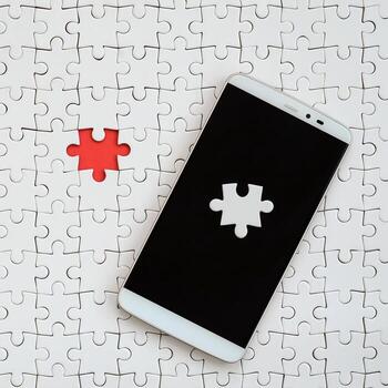 A modern big smartphone with several puzzle elements on the touch screen lies on a white jigsaw puzzle in an assembled state with missing elements photo