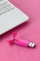 la tarjeta de memoria flash usb de color rosa brillante con un lazo rosa se encuentra sobre una manta de tela suave y peluda de color rosa claro junto a una computadora portátil blanca. diseño clásico de regalo femenino para una tarjeta de memoria foto