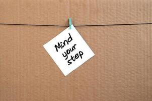 Mind your step. Note is written on a white sticker that hangs with a clothespin on a rope on a background of brown cardboard photo