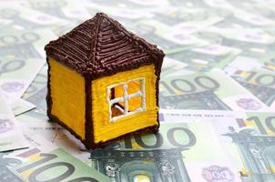 Small toy house is lies on a set of green monetary denominations of 100 euros. A lot of money forms an infinite heap photo