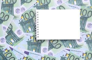 White notebook with clean pages lying on a set of green monetary denominations of 100 euros. A lot of money forms an infinite heap photo