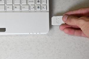 A male hand inserts a white compact SD card into the corresponding input in the side of the white netbook. Man uses modern technologies to store memory and digital data photo