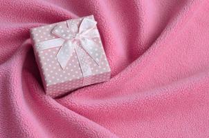 A small gift box in pink with a small bow lies on a blanket of soft and furry light pink fleece fabric with a lot of relief folds. Packing for a gift to your lovely girlfriend photo