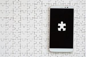 A modern big smartphone with several puzzle elements on the touch screen lies on a white jigsaw puzzle in an assembled state photo