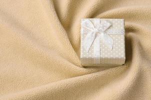 A small gift box in orange with a small bow lies on a blanket of soft and furry light orange fleece fabric with a lot of relief folds. Packing for a gift to your lovely girlfriend photo