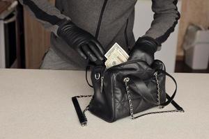 Robber in black outfit and gloves see in opened stolen women bag. The thief takes out US dollar bills from a womans handbag in kitchen photo