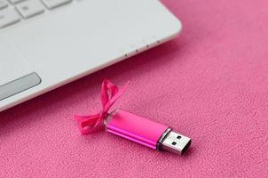 Brilliant pink usb flash memory card with a pink bow lies on a blanket of soft and furry light pink fleece fabric beside to a white laptop. Classic female gift design for a memory card photo