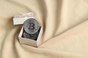 The silver bitcoin lies in a small orange gift box with a small bow on a blanket made of soft and fluffy light orange fleece fabric with a large number of relief folds photo