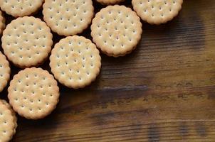 A round sandwich cookie with coconut filling lies in large quantities on a brown wooden surface. Photo of edible treats on a wooden background with copy space