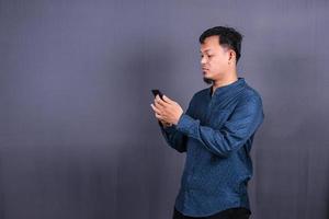 Portrait of unhappy Asian man in blue shirt holding mobile phone with sad expression on face. Isolated on gray background photo