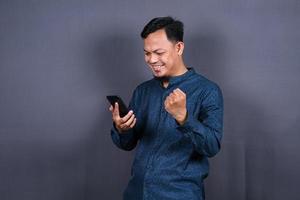 Happy young man in blue shirt posing isolated on gray background studio portrait. People lifestyle concept. Mock up copy space. Hold mobile phone, doing winner gesture photo