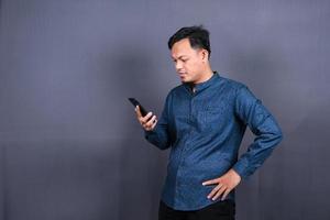 Portrait of unhappy Asian man in blue shirt holding mobile phone with sad expression on face. Isolated on gray background photo