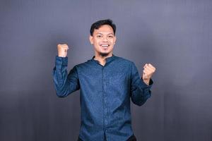 Young handsome man with blue shirt over gray background very happy and excited doing winner gesture with arms raised, smiling and screaming for success. Celebration photo