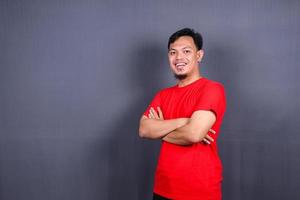 retrato de un atractivo hombre asiático con una camiseta roja de pie con los brazos cruzados aislado en un fondo gris foto