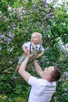 Young father is holding his little son on hands on background of blossom lilac flowers trees. photo