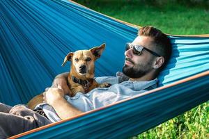 un hombre europeo sonriente y atractivo con gafas de sol descansa en una hamaca con su lindo perrito. foto