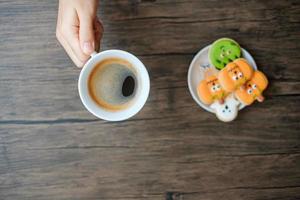 mano sosteniendo una taza de café mientras come divertidas galletas de halloween. feliz día de halloween, truco o amenaza, hola octubre, otoño, concepto tradicional, fiesta y vacaciones foto