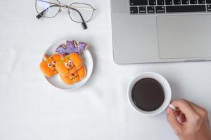 Hand holding coffee cup and eating Halloween Cookies during using computer laptop. Happy Halloween, online shopping, Hello October, fall autumn, Festive, party and holiday concept photo