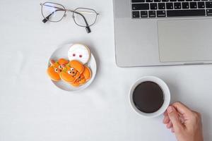 Hand holding coffee cup and eating Halloween Cookies during using computer laptop. Happy Halloween, online shopping, Hello October, fall autumn, Festive, party and holiday concept photo
