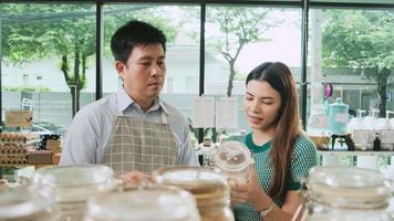 Asian male shopkeeper describes natural organic products to woman customer in refill store, zero-waste and plastic-free grocery, eco environment-friendly, sustainable lifestyles with a reusable shop. video