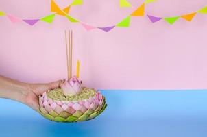 Pink lotus petal krathong that have 3 incense sticks and candle decorates with lotus and crown flower holding by hand for Thailand Loy Krathong festival on pink and blue background. photo