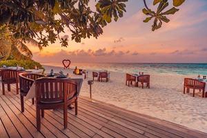 Outdoor restaurant at the beach. Table setting at tropical beach restaurant. Led light candles and wooden tables, chairs under beautiful sunset sky, sea view. Luxury hotel or resort restaurant photo