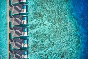 Aerial top view of pool villas, bungalows in Maldives paradise tropical beach. Amazing blue turquoise sea lagoon, ocean bay water. Luxury travel vacation destination. Beautiful sunny aerial landscape photo