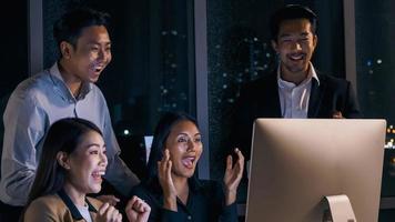 Group of Asian businesspeople looking at computer screen and celebrating success together in the office after working late at night. photo