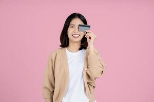 feliz mujer asiática sonriente con ropa informal sosteniendo una tarjeta bancaria y buscando una cámara, aislada de fondo rosa. foto