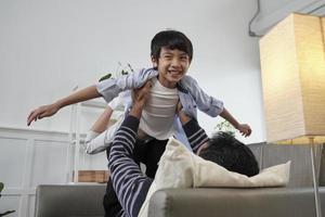 familia tailandesa asiática junta, el padre se acuesta y la diversión juega con el hijo levantando, extendiendo los brazos como un avión volador en el sofá de la sala de estar, tiempos de ocio felices, fin de semana encantador, estilo de vida doméstico de bienestar. foto