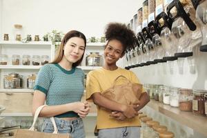 dos clientas miran la cámara y sonríen, compran productos orgánicos en la tienda de recarga con bolsas reutilizables, comestibles sin desperdicio y estilos de vida sostenibles, respetuosos con el medio ambiente y sin plástico. foto