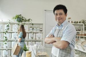 Portrait of Asian male shopkeeper smiles and looks at camera, arms crossed at refill store, natural products, zero-waste grocery, and plastic-free, eco environment-friendly, sustainable lifestyles. photo