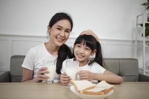 retrato de una familia tailandesa asiática saludable, una hija feliz y una madre joven mirando la cámara, beben leche fresca y pan juntos en la mesa del comedor por la mañana, estilo de vida de comida de desayuno de nutrición de bienestar. foto