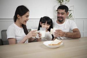 A Healthy Asian Thai family, a little daughter, and young parents drink fresh white milk in glass and bread joy together at a dining table in morning, wellness nutrition home breakfast meal lifestyle. photo