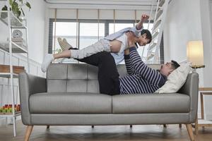 Asian Thai family together, father lies down and fun plays with son by lifting, spread arms like flying airplane on living room sofa, happy leisure times, lovely weekend, wellbeing domestic lifestyle. photo