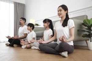 feliz familia tailandesa asiática, padres e hijos se sientan en el piso de la sala de estar, cierran los ojos juntos, meditan tranquilamente en posición de loto y practican yoga saludable, fin de semana de estilo de vida doméstico. foto