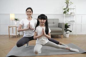 Young Asian Thai mother lovely trains her little daughter to exercise and practices yoga on living room floor together, healthy fitness and wellness, happy domestic home lifestyle on family weekend. photo