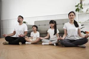 feliz familia tailandesa asiática, padres e hijos se sientan en el piso de la sala de estar, cierran los ojos juntos, meditan tranquilamente en posición de loto y practican yoga saludable, fin de semana de estilo de vida doméstico. foto